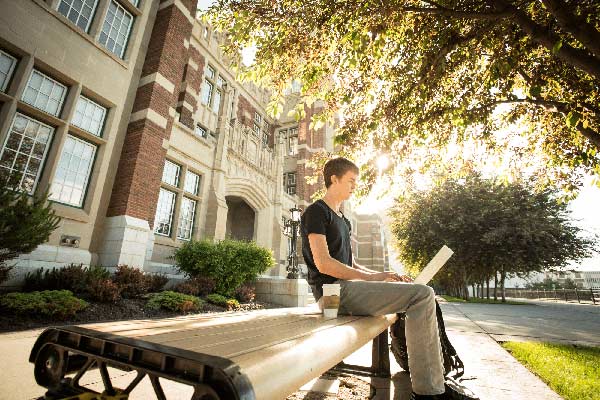 A student works on a laptop, balanced on their lap, while he sits on a bench outside Heritage Hall on a sunny summer day.