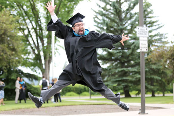 A SAIT graduate wearing their cap and gown jumps up in the air.