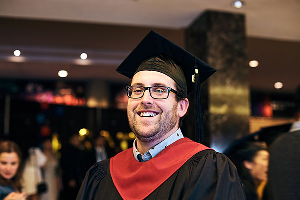 A School of Hospitality and Tourism graduate wearing his cap and gown smiles at the camera.