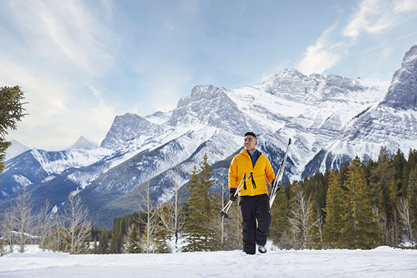 A man wearing a yellow a jacket and black snow pants walks across the snow holding skiis. The Rocky Mountains are behind him.