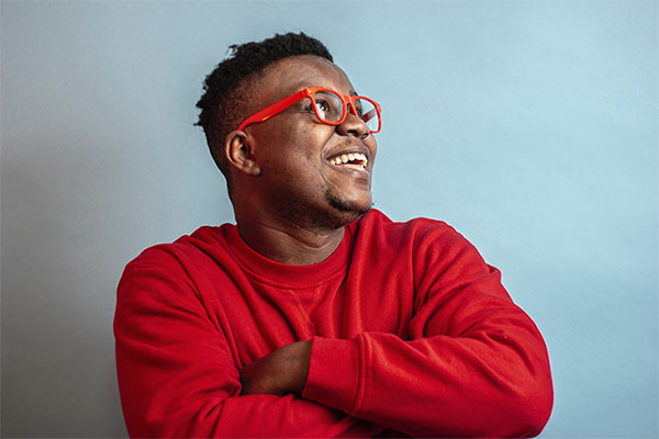 A black man wearing a red long-sleeved shirt crosses his arms. He's wearing matching red glasses, and looking to the right, smiling.