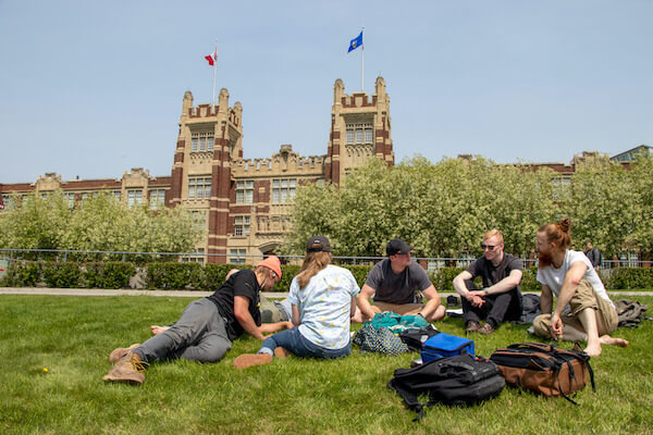 A group of students sit on the Cohos Commons Athletic Field on a summer day. Heritage Hall is seen behind them.