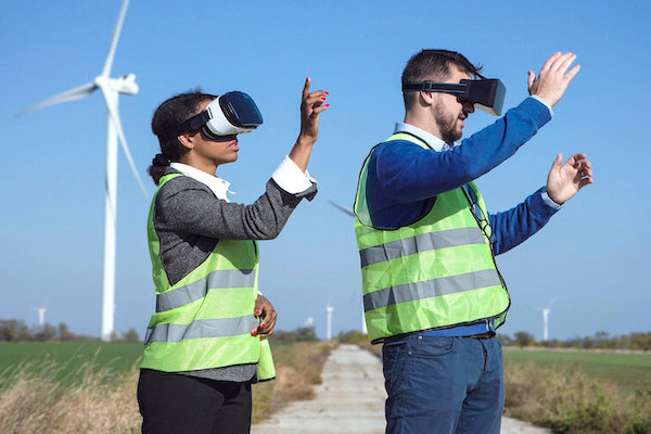 Two people wearing VR classes and safety vests stand in a field with windmills behind them.