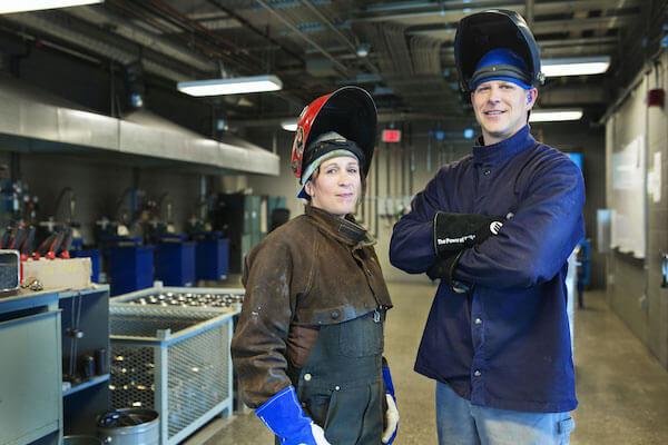 Two people wearing welding helmets look at the camera. 