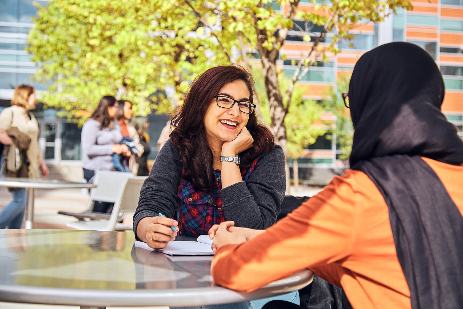 A woman with long, dark brown hair rests her head on her chin while listening to someone else. They are sitting a table outside on SAIT campus during the summer.