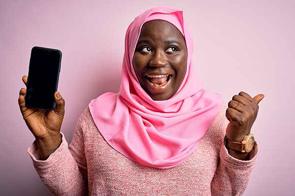 A black woman of colour wearing a pink hijab smiles while pointing and looking to the left, holding her smartphone up in her right hand.