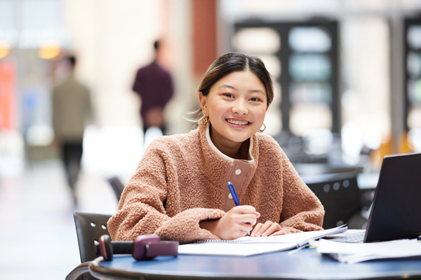 A student looks up from her desk where she's writing on her notebook, and smiles at the camera.