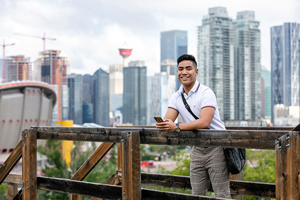 A young man holds his cellphone while leaning up against a wooden railing. Downtown Calgary is seen behind him.