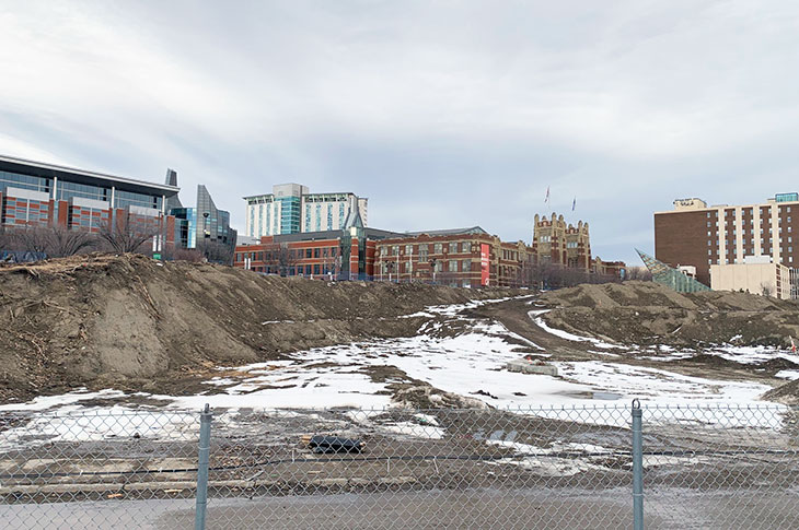 View of empty Campus Centre construction site from the CTrain platform, showing large mounds of dirt with Heritage Hall building in the background.