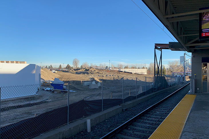 View of Campus Centre construction site from the CTrain platform. Campus buildings, CTrain platform and the Calgary skyline are partially visible. The construction site is mostly dirt mounds.
