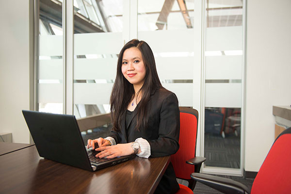 A young woman with long, dark brown hair, wearing a suit jacket, sits at an office desk with a laptop. She's smiling at the camera.