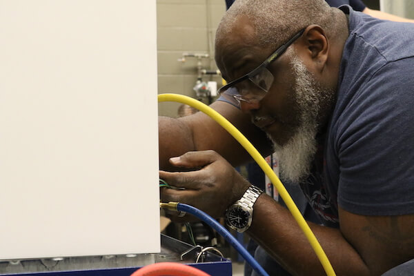 A man with a beard, wearing safety glasses, inspects an appliance's wiring.