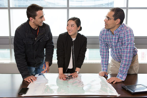 Three peoples analyze a map laid out on a table. 