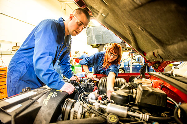 A male and female student wearing safety glasses and coveralls work on a car engine. 