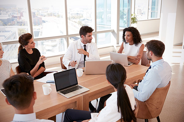 A group of employees in business casual attire sit around a boardroom table having a discussion.