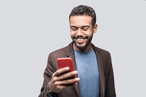 A young man wearing a brown blazer over a blue shirt smiles at his cellphone.