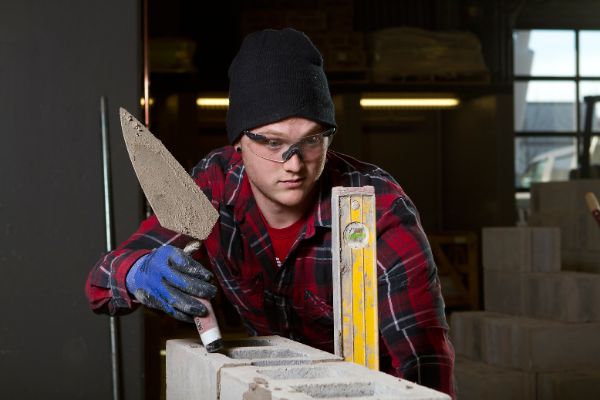 A bricklayer apprentice wearing safety glasses and holding a trowel checks to see if the bricks they are working on are level.