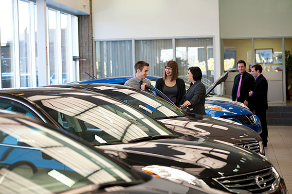 Two women speak with a male salesperson in a dealership, while looking at a Nissan sedan vehicle.