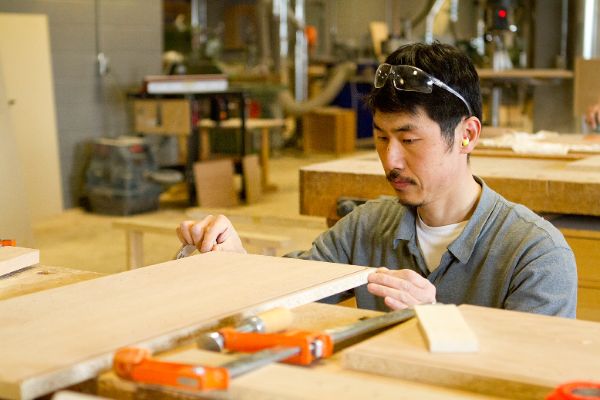 A cabinetmaker apprentice carefully pushes a sheet of wood through a saw.
