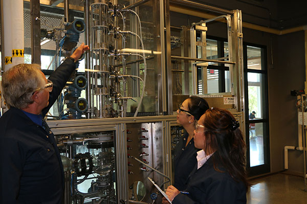Two students take notes while listening to an instructor inside a SAIT chemical lab. They're all wearing safety glasses.