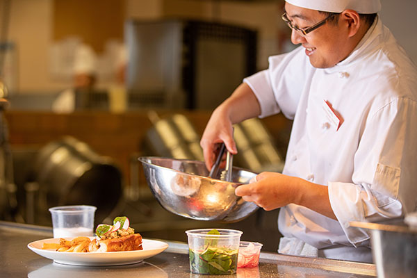 A chef wearing kitchen whites, smiling and seasoning food in a stainless steel bowl.