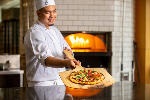 Student holding a pizza on a paddle by a wood buring pizza oven