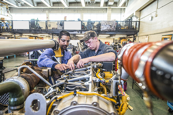 Two students wearing safety glasses and coveralls work together to perform a tune-up on heavy equipment machinery within a SAIT shop lab. 