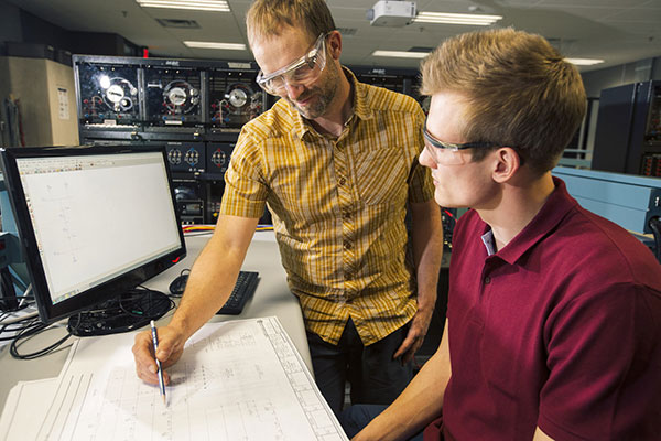 An instructor explains something to an Engineering Design and Drafting student referencing some engineering drawings. They are both wearing safety glasses.