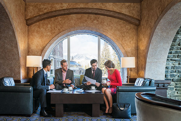 Three men and a woman wearing business formal attire sit around a low table having a discussion inside a luxury hotel in the mountains.