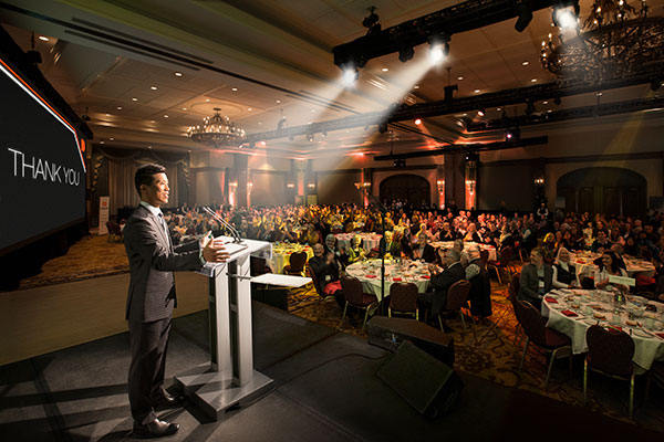 An emcee speaking from the stand facing a crowd of people seated in tables