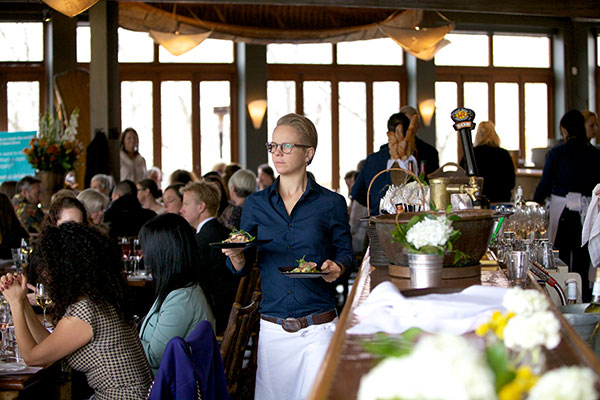 A blonde woman with her hair up in a bun carries a tray of glasses, walking through a crowded restaurant. 