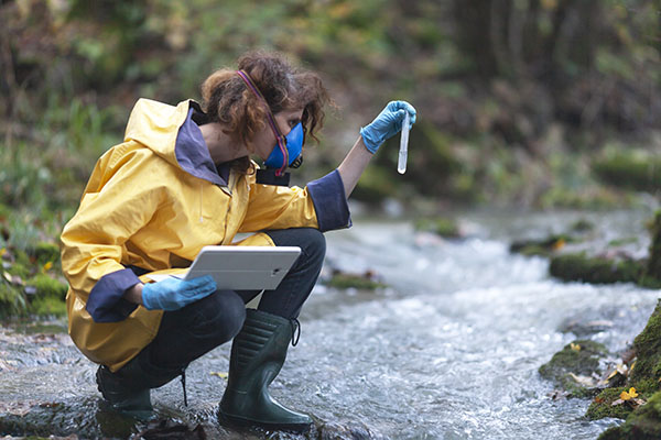 A student holding a tool while crouching down on a shallow part of a river and holding an ipad on the other hand