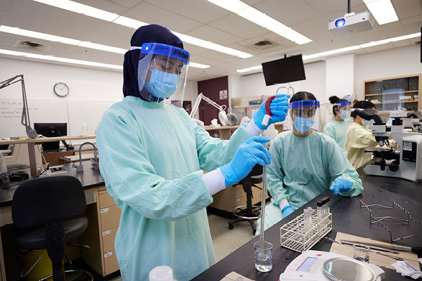 Students wearing scrubs, face masks and face shields work in a medical lab classroom.