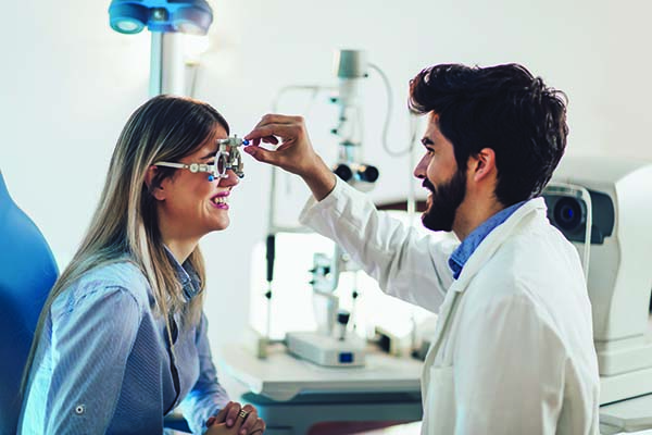 An optician student wearing a lab coat performs visual measurements on a smiling patient.