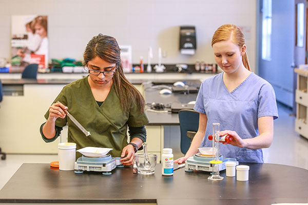 Two young women wearing scrubs measure out prescription medication in a SAIT lab.