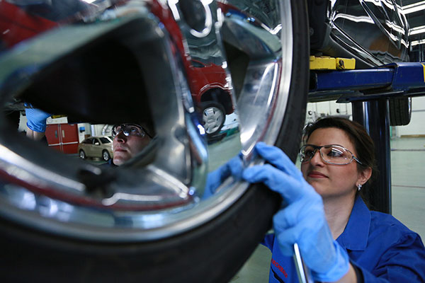 A young woman wearing safety glasses and coveralls checks the tire pressure of a vehicle on a lift.