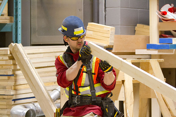 A young man wearing ear plugs, a hard hat, safety glasses and a safety vest inspects a 2x4 plank of wood. Piles of 2x4s are seen all around him.