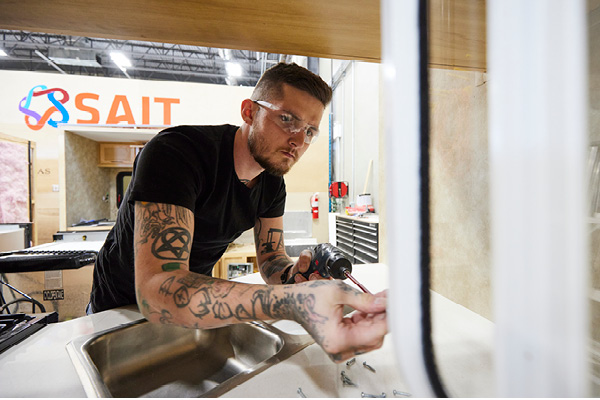 An RV service technician apprentice works inside the storage compartment of a travel trailer.