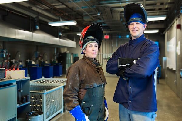 Two people wearing welding shields on top of their heads look at the camera