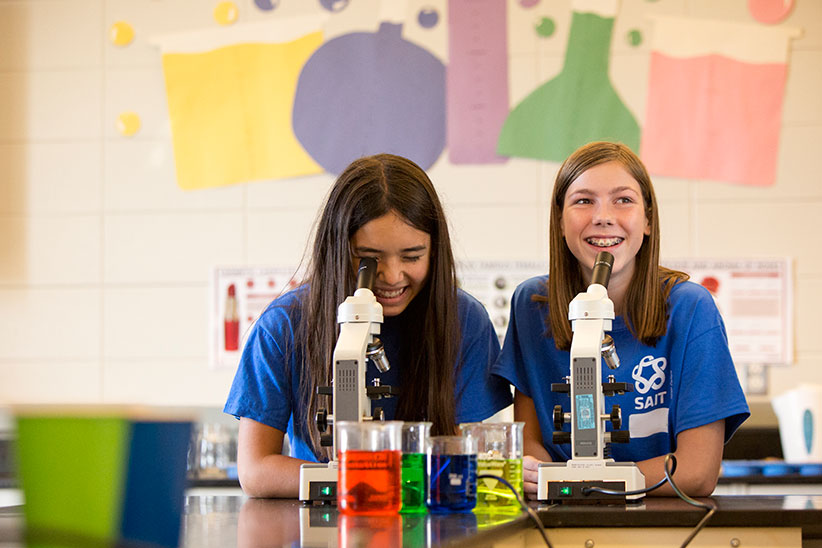 Two SAIT campers look through microscopes. Behind them, the wall is decorated with the shape of beakers. In front of them are four glass jars, each filled with a coloured liquid.