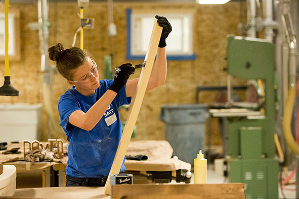 A young woman lacquers a wood 2 by 4. She's wearing protective gloves and safety glasses.