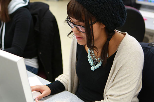 A young woman with glasses, wearing a buret and cardigan, works on a computer.
