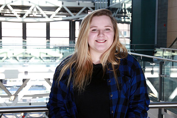 A young high school student with long, blonde hair, wearing a button down blue plaid shirt smiles at the camera. She is standing in the Aldred Centre on SAIT campus.