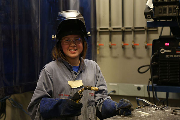 A young woman wearing full welding PPE has her helmet up and smiles for the camera.